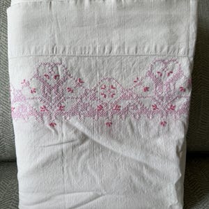 May include: Folded white sheet with a decorative pink embroidered border. The embroidery features a repeating floral and geometric pattern. The sheet is made of a soft, textured fabric and is neatly folded.