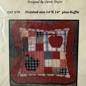 May include: A patchwork pillow with a red ruffle, featuring apple designs. The pillow's finished size is 14" x 14". The design includes squares of red, white, and blue fabrics. The text "APPLE PILLOW" is at the top.