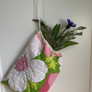 May include: A pink and green floral fabric bag with a drawstring closure, hanging from a hook. The bag is filled with a bouquet of purple flowers and green foliage.