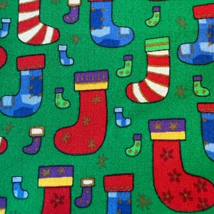 May include: A green fabric featuring a repeating pattern of Christmas stockings in various sizes and designs. The stockings are colored in red, blue, and purple, with some featuring stripes, star motifs, or solid blocks of color against the green background.