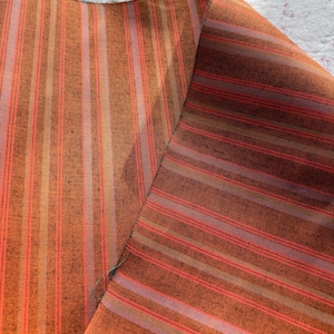 May include: Two pieces of fabric with vertical stripes in shades of orange, red, and light pink. The fabric has a textured appearance, with the stripes varying in width. The fabric is folded over, revealing the reverse side.