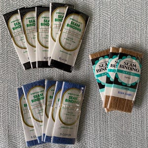 May include: Assortment of stretch lace seam binding in black, blue, and brown. The packages are labeled with the brand name "J & P Coats" and the length of the binding in yards. The colors are black, blue, and camel.