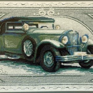 May include: Cross-stitch artwork of a vintage green car with a black top, set within a decorative frame. The car features detailed headlights and spoked wheels, rendered in shades of green, blue, and gray.
