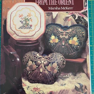 May include: A book titled "Needlework Treasures From The Orient" by Marsha McKerr, featuring a close-up of three embroidered cushions with floral and butterfly designs.