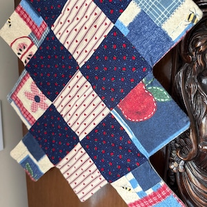 May include: A quilted, patchwork item with a variety of patterns. The design includes squares of navy blue with red dots, cream with red hearts, and blue plaid. There are also squares with red apples and a bee.