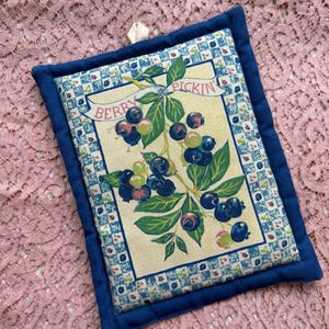 May include: A rectangular quilted pot holder with a blue border and a cream-colored center. The center features a vintage illustration of blueberries and the text "Berry Pickin". A small loop is attached for hanging.