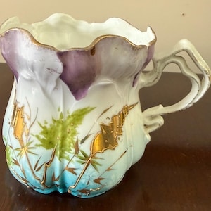 Peut inclure: Tasse à thé ancienne en porcelaine avec une anse unique et un motif floral. La tasse présente une base blanche avec un motif floral bleu et vert, rehaussé d'une bordure dorée et d'accents violets. La poignée a un design orné et ouvert.