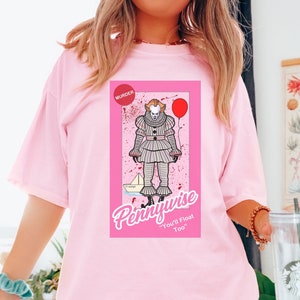 May include: A light pink t-shirt featuring a graphic print of the character Pennywise in a clown costume, accompanied by a red balloon, a paper boat, and red splatters. The text reads "MURDER", "Pennywise", and "You'll Float Too" in white and red.