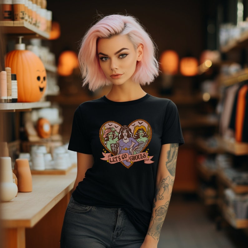 May include: Black t-shirt featuring a heart-shaped graphic with three cartoon ghouls and the text "Let's Go Ghouls." The design includes bats and a banner. The shirt is a Halloween-themed apparel item.
