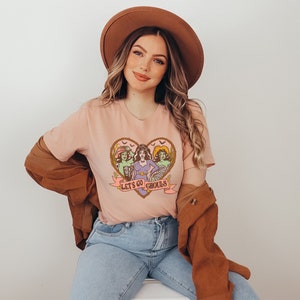 May include: A light pink t-shirt featuring a heart-shaped graphic with three cartoon ghouls and the text "Let's Go Ghouls." The graphic is in shades of brown, green, purple, and pink. The model is wearing a brown hat, a brown jacket, and blue jeans.