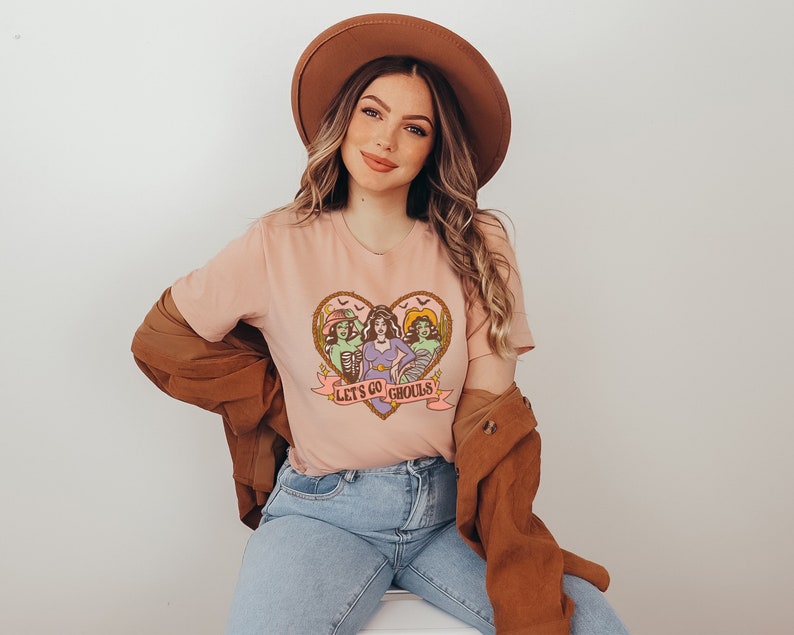 May include: A light pink t-shirt featuring a heart-shaped graphic with three cartoon ghouls and the text "Let's Go Ghouls." The graphic is in shades of brown, green, purple, and pink. The model is wearing a brown hat, a brown jacket, and blue jeans.