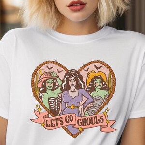May include: White t-shirt featuring a heart-shaped graphic with three cartoon ghouls. The ghouls are in various costumes, with the text "LET'S GO GHOULS" on a banner below. The design includes bats, cacti, and a crescent moon.