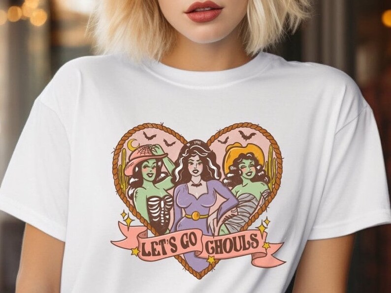 May include: White t-shirt featuring a heart-shaped graphic with three cartoon ghouls. The ghouls are in various costumes, with the text "LET'S GO GHOULS" on a banner below. The design includes bats, cacti, and a crescent moon.