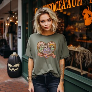 May include: Olive green t-shirt featuring a heart-shaped graphic with three cartoon ghouls and the text "Let's Go Ghouls" on a banner. The design has a vintage, retro style. The model is wearing the shirt outdoors.