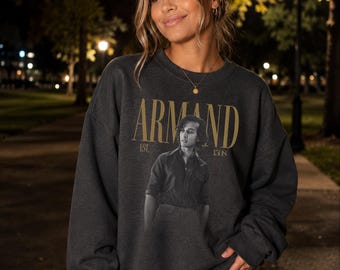 Puede incluir: Una sudadera de cuello redondo en color gris carbón oscuro con un estampado gráfico de una persona con camisa abotonada. El texto ARMAND está impreso en letras doradas grandes sobre la figura, con EST. 1508 en letras doradas más pequeñas.