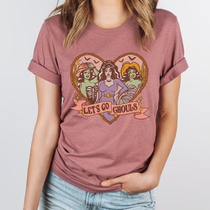 May include: Mauve-colored t-shirt featuring a heart-shaped design with three cartoon ghouls. The ghouls are in a heart-shaped frame with the text "LET'S GO GHOULS" on a banner. The shirt has rolled-up sleeves.