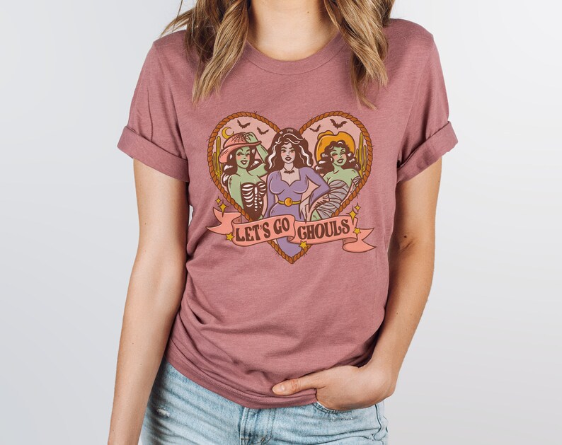 May include: Mauve-colored t-shirt featuring a heart-shaped design with three cartoon ghouls. The ghouls are in a heart-shaped frame with the text "LET'S GO GHOULS" on a banner. The shirt has rolled-up sleeves.