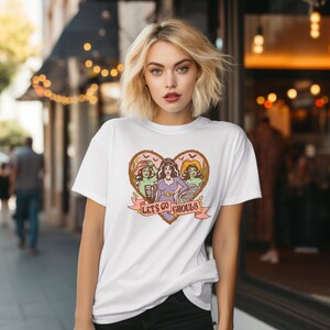 May include: White t-shirt featuring a heart-shaped graphic with three stylized female figures dressed as Halloween ghouls. The phrase "Let's Go Ghouls" is printed on a banner below the heart. The design has a vintage, cartoonish style.