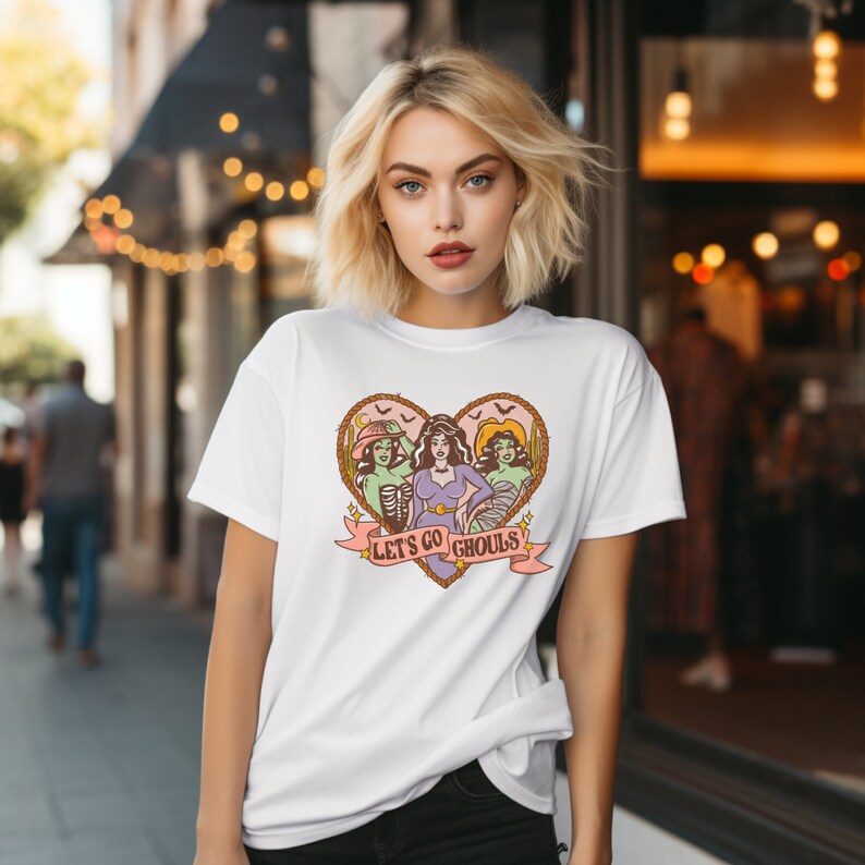 May include: White t-shirt featuring a heart-shaped graphic with three stylized female figures dressed as Halloween ghouls. The phrase "Let's Go Ghouls" is printed on a banner below the heart. The design has a vintage, cartoonish style.