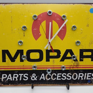 May include: A yellow MOPAR clock with a distressed finish. The clock face features the MOPAR logo in red, a red and white clock hand, and the words "PARTS & ACCESSORIES". The clock is mounted with metal nuts.