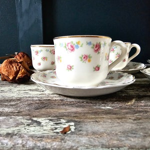 May include: Set of four white teacups and saucers with a floral pattern in pink, blue, and yellow. The cups have gold rims and the saucers have a scalloped edge.