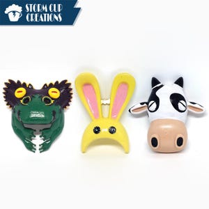 May include: Three whimsical animal-themed magnets: a green frog with yellow eyes, a yellow bunny with pink ears, and a black and white cow. The magnets are brightly colored and have a cartoonish design.