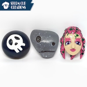 May include: Three painted decorative stones. One is dark blue with a white skull, another is gray with speckles and a face, and the third is a face with pink hair and green vines. The Storm Cup Creations logo is in the upper left corner.