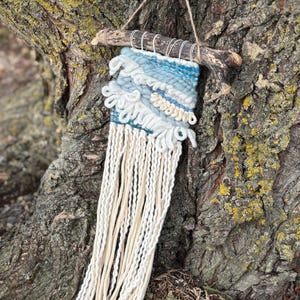 May include: A handcrafted wall hanging featuring a woven design in shades of blue and white, suspended from a natural wooden branch. The textile art piece has a textured surface with long, cream-colored fringe.