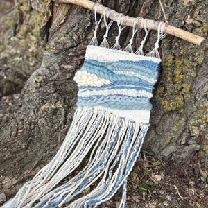 May include: A woven wall hanging with a natural wooden branch and twine. The textile features a blend of white, light blue, and medium blue yarns, creating a textured, layered design. Long, braided fringe hangs from the bottom.