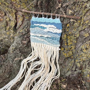May include: Handwoven wall hanging featuring a landscape design in shades of blue, white, and cream. The textile art piece is suspended from a natural wooden branch with a jute string. The bottom of the piece has long, cream-colored fringe.