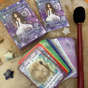 May include: An oracle deck with the title "Soul Vibrations" and a matching box. The deck features colorful cards with illustrations and text, along with decorative star and moon-shaped stones. A drum mallet and a deck of cards are also present.