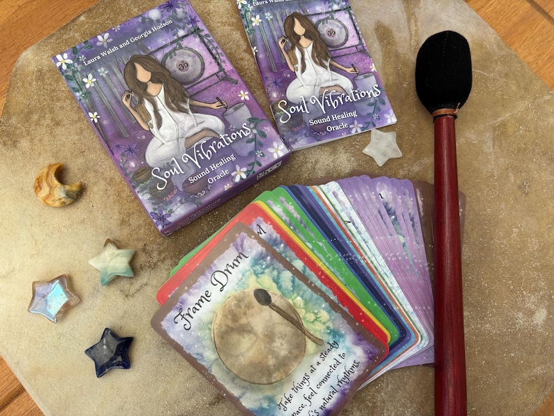 May include: An oracle deck with the title "Soul Vibrations" and a matching box. The deck features colorful cards with illustrations and text, along with decorative star and moon-shaped stones. A drum mallet and a deck of cards are also present.