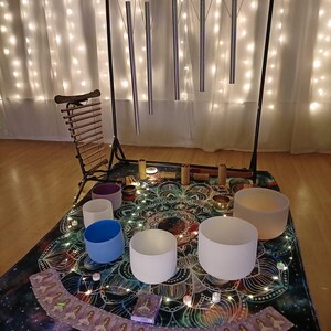May include: A sound healing setup featuring a black metal stand with hanging chimes, a xylophone, and several crystal singing bowls in various colors. The setup is on a patterned mat with fairy lights.