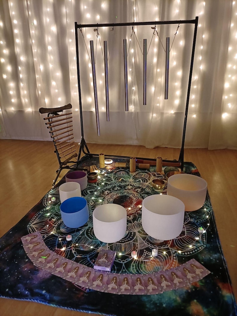 May include: A sound healing setup featuring a black metal stand with hanging chimes, a xylophone, and several crystal singing bowls in various colors. The setup is on a patterned mat with fairy lights.