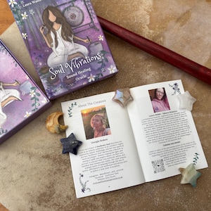 May include: A purple oracle card box and deck with the title "Soul Vibrations" and artwork of a woman. An open booklet with creator photos and star-shaped crystals are also visible. A wooden drumstick rests on the surface.