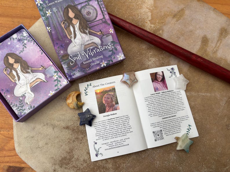 May include: A purple oracle card box and deck with the title "Soul Vibrations" and artwork of a woman. An open booklet with creator photos and star-shaped crystals are also visible. A wooden drumstick rests on the surface.