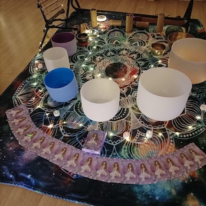 May include: A collection of sound healing instruments, including crystal singing bowls, chimes, and a set of oracle cards, arranged on a patterned mat. The bowls are various colors, and fairy lights are scattered around the setup.