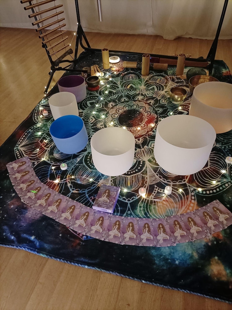 May include: A collection of sound healing instruments, including crystal singing bowls, chimes, and a set of oracle cards, arranged on a patterned mat. The bowls are various colors, and fairy lights are scattered around the setup.