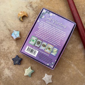 May include: A purple box with the text "Soul Vibrations Sound Healing Oracle" is surrounded by star-shaped and crescent-shaped stones. A wooden drumstick with a black head rests on the right side. The box contains 44 cards and a guidebook.