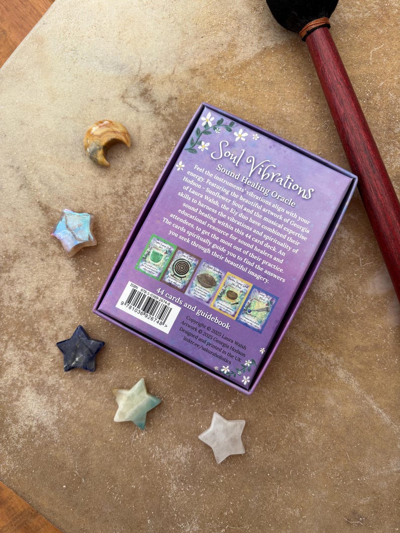 May include: A purple box with the text "Soul Vibrations Sound Healing Oracle" is surrounded by star-shaped and crescent-shaped stones. A wooden drumstick with a black head rests on the right side. The box contains 44 cards and a guidebook.