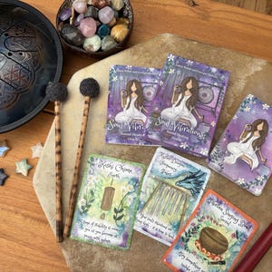 May include: An assortment of spiritual tools and accessories, including a steel tongue drum, mallets, oracle cards, and a bowl of crystals. The cards feature illustrations and text, with a focus on sound healing and spirituality.