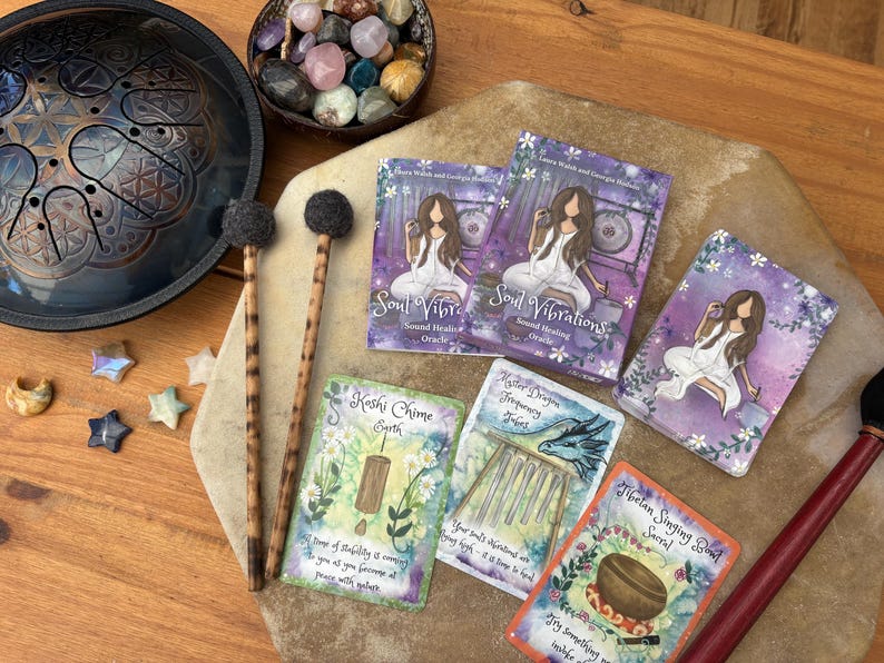 May include: An assortment of spiritual tools and accessories, including a steel tongue drum, mallets, oracle cards, and a bowl of crystals. The cards feature illustrations and text, with a focus on sound healing and spirituality.