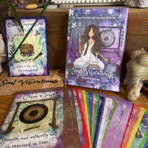 May include: A Soul Vibrations Sound Healing Oracle deck with a purple cover featuring an illustration of a woman and a gong. The deck includes cards with images of singing bowls and gongs. The text on the cards includes "Tibetan Singing Bowl" and "Chau Gong".