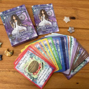 May include: Two decks of "Soul Vibrations" oracle cards with a woman illustration, and a fan of colorful cards. The cards are on a wooden surface with star and crescent-shaped decorative stones.