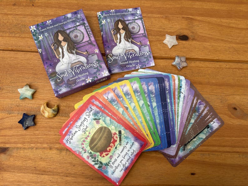 May include: Two decks of "Soul Vibrations" oracle cards with a woman illustration, and a fan of colorful cards. The cards are on a wooden surface with star and crescent-shaped decorative stones.