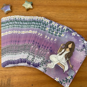May include: A deck of tarot cards with a purple and white floral design. The cards feature an illustration of a woman in a white dress. Decorative star and moon-shaped crystals are scattered around the deck on a wooden surface.