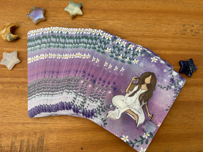 May include: A deck of tarot cards with a purple and white floral design. The cards feature an illustration of a woman in a white dress. Decorative star and moon-shaped crystals are scattered around the deck on a wooden surface.
