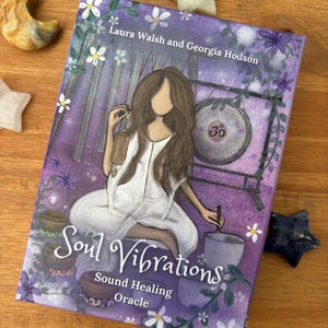 May include: A book titled "Soul Vibrations" with a purple cover featuring an illustration of a woman in white. The book is surrounded by star-shaped and crescent-shaped crystals. The text "Sound Healing Oracle" is also visible.