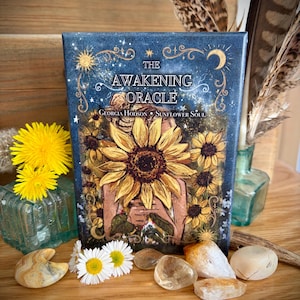 May include: A rectangular oracle card deck box titled The Awakening Oracle by Georgia Hodson Sunflower Soul. The box features a dark blue background with gold celestial motifs and a central illustration of a person holding a large yellow sunflower.