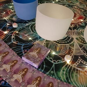 May include: A collection of frosted glass singing bowls in various colors, including white, blue, and purple, arranged on a patterned cloth. A deck of "Soul Vibrations" oracle cards and small candles are also present, suggesting a spiritual or wellness theme.